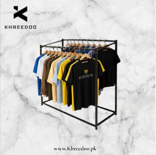 A black metal clothing rack holding multiple t-shirts in assorted colors, with a black t-shirt in the front featuring a yellow graphic and the word 'WARRIOR'. The image is set against a white marble background with the brand name 'KHREEDOO' in the top left and a website URL at the bottom.