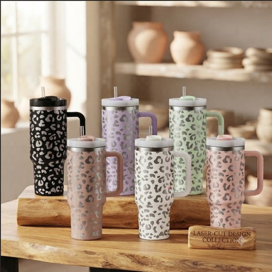 Six insulated travel tumblers with handles and straws are arranged on a wooden table in a softly lit interior. The tumblers have a repeating leopard-style spot pattern in different colorways: black and white, lavender and white, mint green and white, blush pink and white, and taupe and white.
